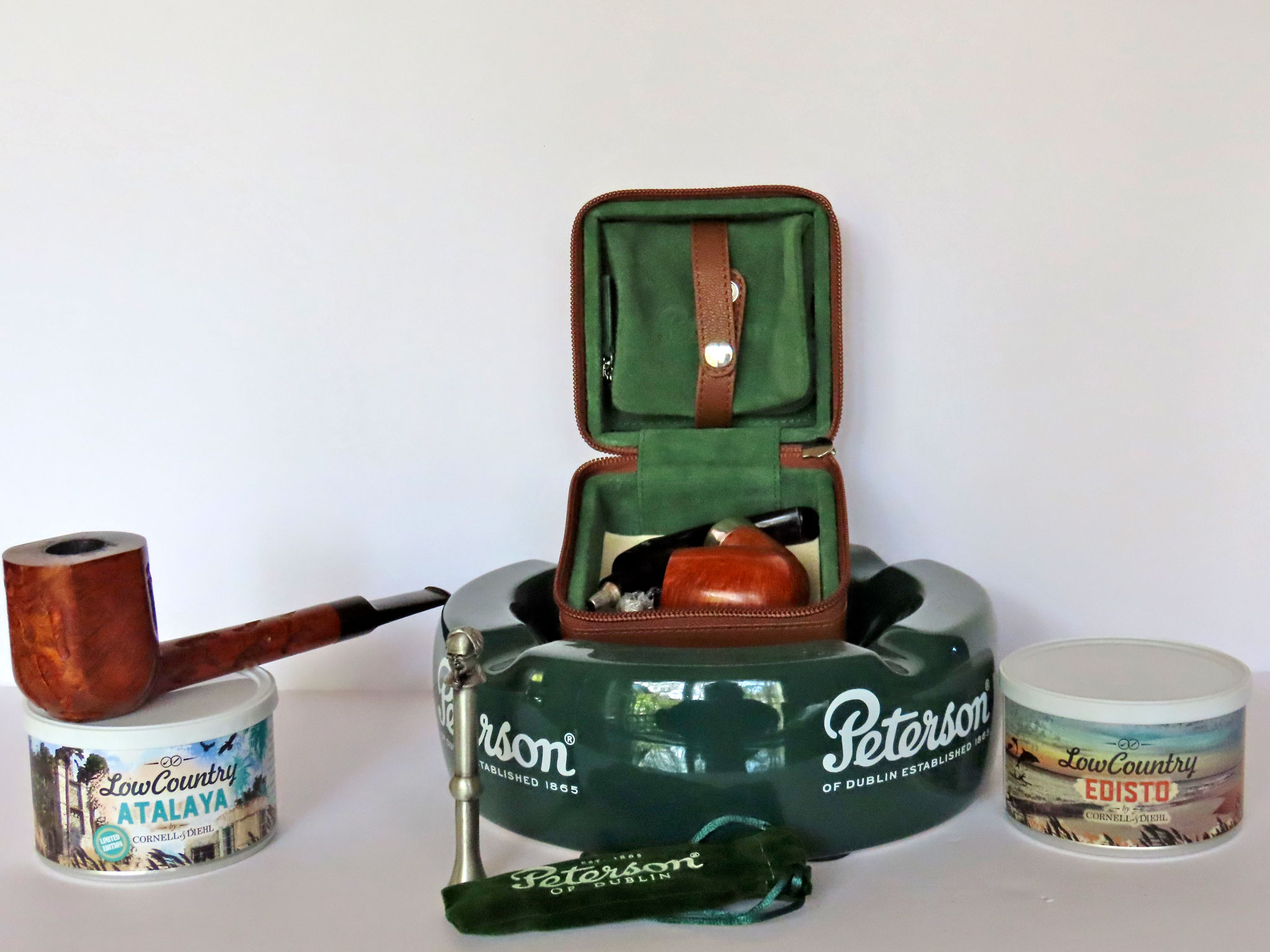 From L-R: The Rich Lewis Custom Bill NASPC 2014 POY atop a fresh tin of Low County Atalaya a ready-rubbed blend combining Red and Bright Virginias from 2019 with C&D's proprietary Red Virginia Cavendish.Peterson’s Grafton System Pipe Travel Case specifically designed for Peterson System pipes, with matching leather pipe stand and a green suede tobacco pouch. Front: Peterson's Thinking Man pewter tamper, attributed to Peterson’s 1905 slogan, "The Thinking Man Smokes a Peterson Pipe." Far right: Fresh tin of C&D Low Country Edisto blend, Red Virginias pressed and sliced into flakes. Photo by Fred Brown